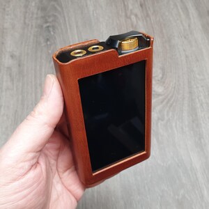Puede incluir: Un reproductor de audio port&aacute;til marr&oacute;n con una pantalla negra y detalles dorados. El dispositivo tiene una textura similar a la madera y un bot&oacute;n dorado. El reproductor se sostiene en una mano.