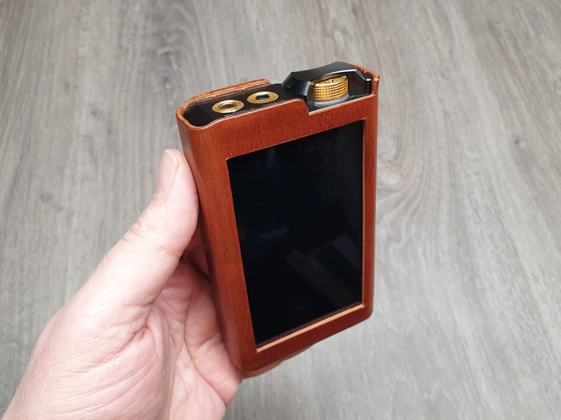 Puede incluir: Un reproductor de audio port&aacute;til marr&oacute;n con una pantalla negra y detalles dorados. El dispositivo tiene una textura similar a la madera y un bot&oacute;n dorado. El reproductor se sostiene en una mano.