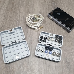 Könnte beinhalten: Ein digitaler Audioplayer mit schwarzem Bildschirm, ein Paar kabelgebundene In-Ear-Kopfhörer mit geflochtenem silbernem Kabel und zwei graue Kunststoff-Aufbewahrungsboxen mit verschiedenen schwarzen, weißen, blauen und grauen Silikon-Ohrstöpseln.