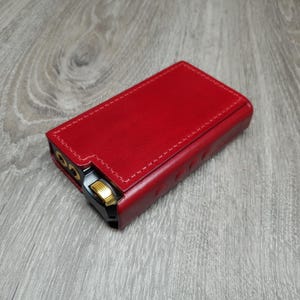 Puede incluir: Una funda de cuero roja para un reproductor de música portátil. La funda tiene un borde cosido y detalles dorados.