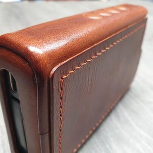 Puede incluir: Primer plano de una funda de cuero marr&oacute;n con costuras visibles. La funda tiene forma rectangular con bordes redondeados y una superficie lisa y pulida. El cuero tiene un tono rico y c&aacute;lido.