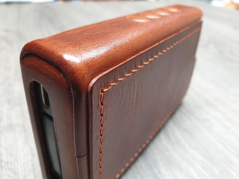 Puede incluir: Primer plano de una funda de cuero marr&oacute;n con costuras visibles. La funda tiene forma rectangular con bordes redondeados y una superficie lisa y pulida. El cuero tiene un tono rico y c&aacute;lido.