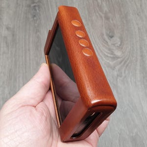 Puede incluir: Una funda de cuero marr&oacute;n para un reproductor multimedia port&aacute;til. La funda tiene forma rectangular con bordes redondeados y cuatro peque&ntilde;os botones ovalados en el lateral. La funda se sostiene en una mano, mostrando la pantalla del dispositivo.