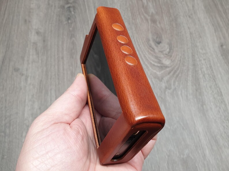 Puede incluir: Una funda de cuero marr&oacute;n para un reproductor multimedia port&aacute;til. La funda tiene forma rectangular con bordes redondeados y cuatro peque&ntilde;os botones ovalados en el lateral. La funda se sostiene en una mano, mostrando la pantalla del dispositivo.