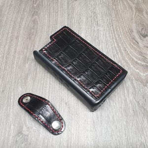 Puede incluir: Juego de funda de teléfono y llavero de cuero negro. La funda del teléfono tiene una textura de piel de cocodrilo con costuras rojas. El llavero es un pequeño accesorio a juego con cierre de botón a presión.