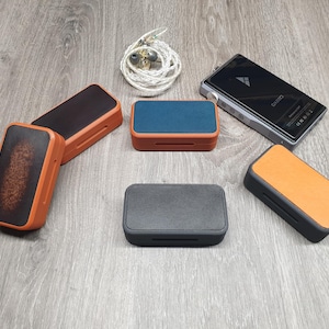Op de afbeelding: Een verzameling rechthoekige opbergdoosjes in diverse kleuren zoals oranje, zwart en blauw, samen met een zwarte digitale audiospeler met een zilveren draaiknop en een opgerold paar zilveren bedrade in-ear hoofdtelefoons met doorzichtige dopjes.