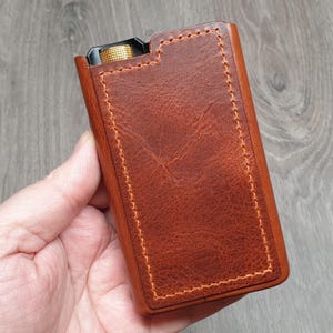 Puede incluir: Una funda de cuero marr&oacute;n con costuras naranjas. La funda tiene forma rectangular y una superficie texturizada. Se ve un bot&oacute;n dorado en la parte superior. La funda est&aacute; sujeta en la mano de una persona.