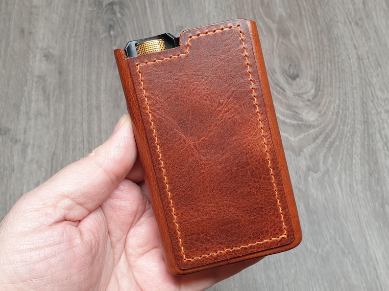 Puede incluir: Una funda de cuero marr&oacute;n con costuras naranjas. La funda tiene forma rectangular y una superficie texturizada. Se ve un bot&oacute;n dorado en la parte superior. La funda est&aacute; sujeta en la mano de una persona.