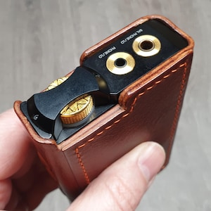 Puede incluir: Una funda de cuero marr&oacute;n con detalles dorados encierra un dispositivo de audio negro. El dispositivo tiene conectores para auriculares dorados y un control de volumen. La funda est&aacute; cosida con hilo naranja.