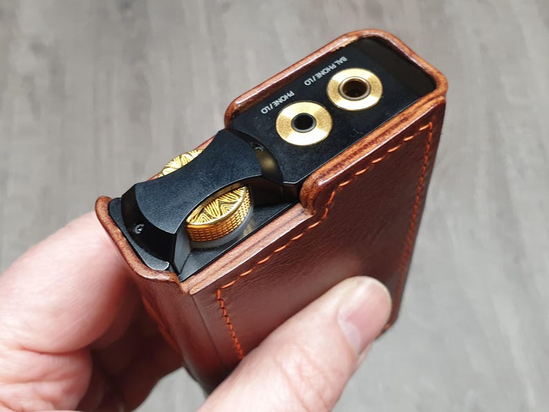 Puede incluir: Una funda de cuero marr&oacute;n con detalles dorados encierra un dispositivo de audio negro. El dispositivo tiene conectores para auriculares dorados y un control de volumen. La funda est&aacute; cosida con hilo naranja.