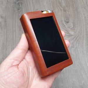 Puede incluir: Una funda de cuero marr&oacute;n con una pantalla negra, probablemente para un dispositivo electr&oacute;nico. La funda rectangular se sostiene en una mano. El fondo es una superficie de madera. El dispositivo tiene un acento met&aacute;lico en la parte superior.