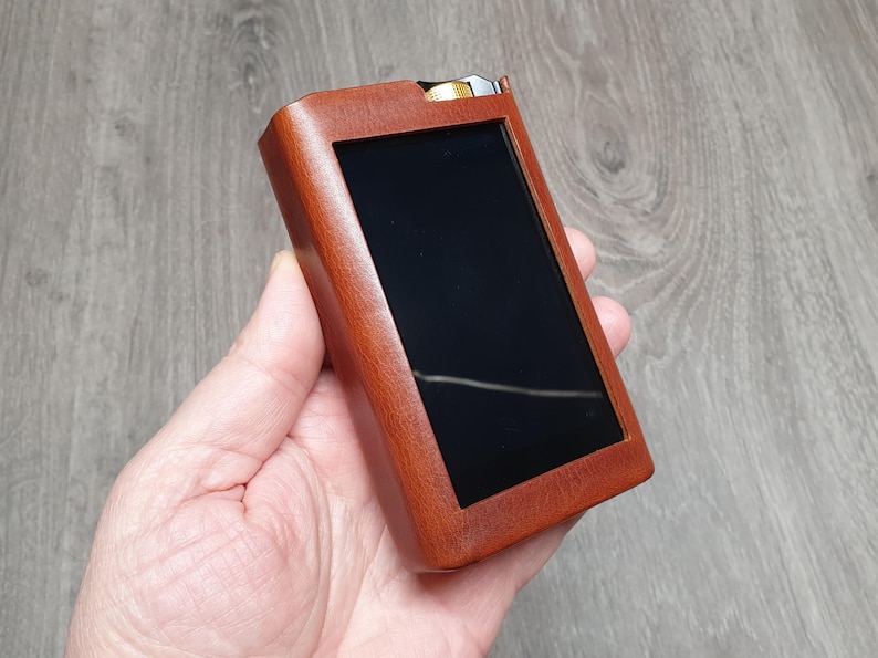 Puede incluir: Una funda de cuero marr&oacute;n con una pantalla negra, probablemente para un dispositivo electr&oacute;nico. La funda rectangular se sostiene en una mano. El fondo es una superficie de madera. El dispositivo tiene un acento met&aacute;lico en la parte superior.