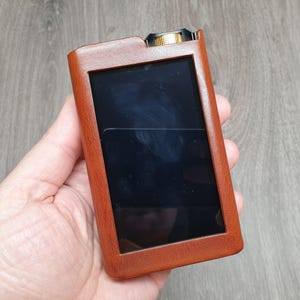 Puede incluir: Una funda de cuero marr&oacute;n para un reproductor multimedia port&aacute;til. La funda tiene forma rectangular con una pantalla negra y un bot&oacute;n dorado en la parte superior. El dispositivo se sostiene en una mano.
