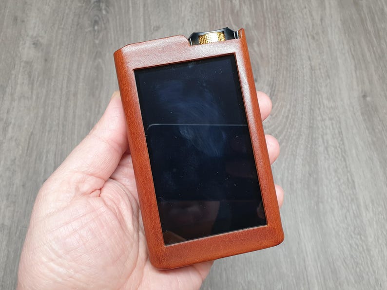 Puede incluir: Una funda de cuero marr&oacute;n para un reproductor multimedia port&aacute;til. La funda tiene forma rectangular con una pantalla negra y un bot&oacute;n dorado en la parte superior. El dispositivo se sostiene en una mano.