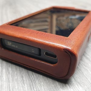 Puede incluir: Una funda de cuero marr&oacute;n para un reproductor de audio digital. La funda tiene forma rectangular con un protector de pantalla transparente y una ranura para una tarjeta SD. La funda est&aacute; hecha de cuero texturizado.