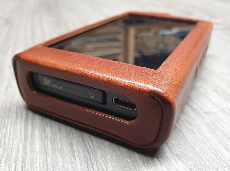 Puede incluir: Una funda de cuero marr&oacute;n para un reproductor de audio digital. La funda tiene forma rectangular con un protector de pantalla transparente y una ranura para una tarjeta SD. La funda est&aacute; hecha de cuero texturizado.