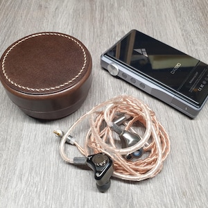 Puede incluir: Un estuche de cuero marrón con costuras blancas, un reproductor de audio digital plateado y negro, y auriculares internos con un cable color cobre se muestran sobre una superficie de madera. El reproductor de audio tiene el texto "DX320".