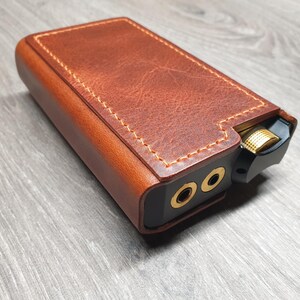 Puede incluir: Una funda de cuero marr&oacute;n con costuras doradas encierra un dispositivo de audio negro. El dispositivo tiene dos conectores de audio dorados y una perilla de control de volumen dorada. La funda tiene una apariencia texturizada.
