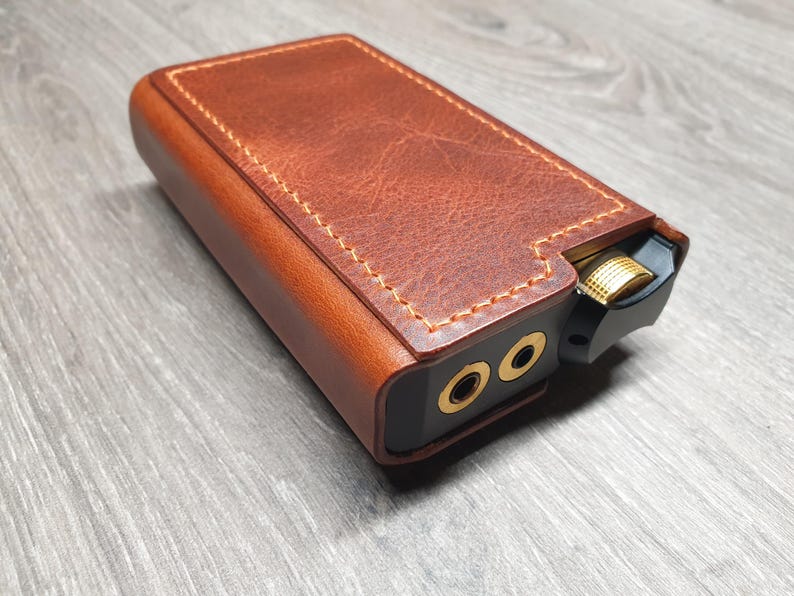 Puede incluir: Una funda de cuero marr&oacute;n con costuras doradas encierra un dispositivo de audio negro. El dispositivo tiene dos conectores de audio dorados y una perilla de control de volumen dorada. La funda tiene una apariencia texturizada.