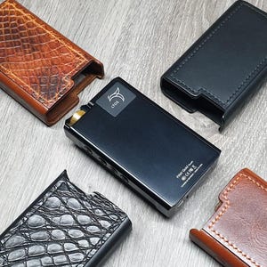 Puede incluir: Un reproductor de música portátil negro y dorado con una pantalla que dice "iBasso DX300" y "FiiO Q5" en la parte posterior. El reproductor está rodeado por cuatro fundas de cuero diferentes, dos marrones y dos negras.