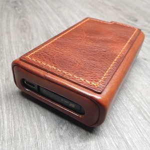 Puede incluir: Una funda rectangular de cuero marr&oacute;n con un borde cosido de color marr&oacute;n claro. La funda tiene una superficie lisa y texturizada y una ranura en un lado. La funda est&aacute; probablemente dise&ntilde;ada para guardar un peque&ntilde;o dispositivo electr&oacute;nico.