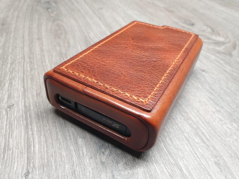 Puede incluir: Una funda rectangular de cuero marr&oacute;n con un borde cosido de color marr&oacute;n claro. La funda tiene una superficie lisa y texturizada y una ranura en un lado. La funda est&aacute; probablemente dise&ntilde;ada para guardar un peque&ntilde;o dispositivo electr&oacute;nico.