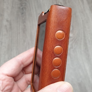 Puede incluir: Primer plano de un reproductor de audio digital de cuero marr&oacute;n. El dispositivo tiene una pantalla rectangular y cuatro botones circulares en el lateral. El cuero tiene un aspecto texturizado.