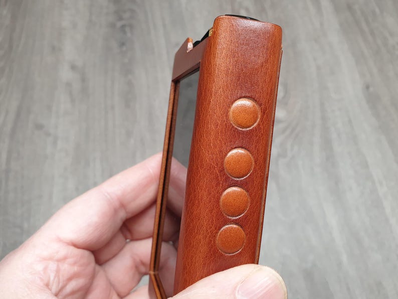 Puede incluir: Primer plano de un reproductor de audio digital de cuero marr&oacute;n. El dispositivo tiene una pantalla rectangular y cuatro botones circulares en el lateral. El cuero tiene un aspecto texturizado.
