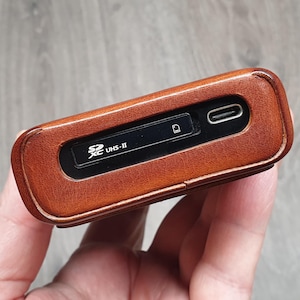 Puede incluir: Un lector de tarjetas SD de cuero marr&oacute;n con un puerto USB-C. El dispositivo tiene un panel negro con el texto "SD XC UHS-II" y un s&iacute;mbolo de ranura para tarjetas. El lector se sostiene en una mano, con una superficie de madera en el fondo.