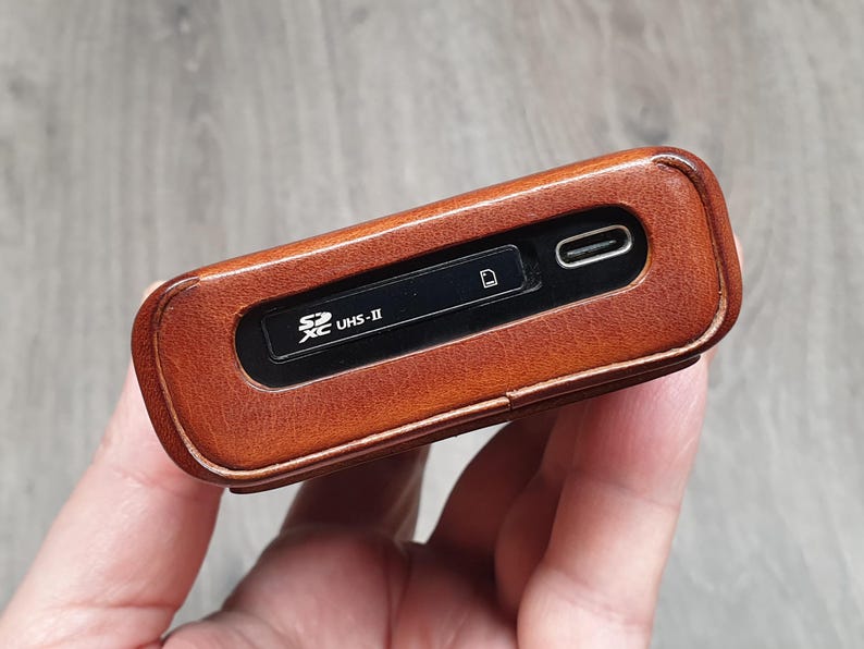 Puede incluir: Un lector de tarjetas SD de cuero marr&oacute;n con un puerto USB-C. El dispositivo tiene un panel negro con el texto "SD XC UHS-II" y un s&iacute;mbolo de ranura para tarjetas. El lector se sostiene en una mano, con una superficie de madera en el fondo.