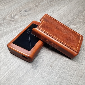 Puede incluir: Una funda y tapa de cuero marr&oacute;n para un reproductor de m&uacute;sica port&aacute;til. La funda est&aacute; abierta, revelando la pantalla del reproductor. El cuero tiene un tono rico y c&aacute;lido, con costuras visibles. El reproductor tiene una pantalla negra y un puerto circular.