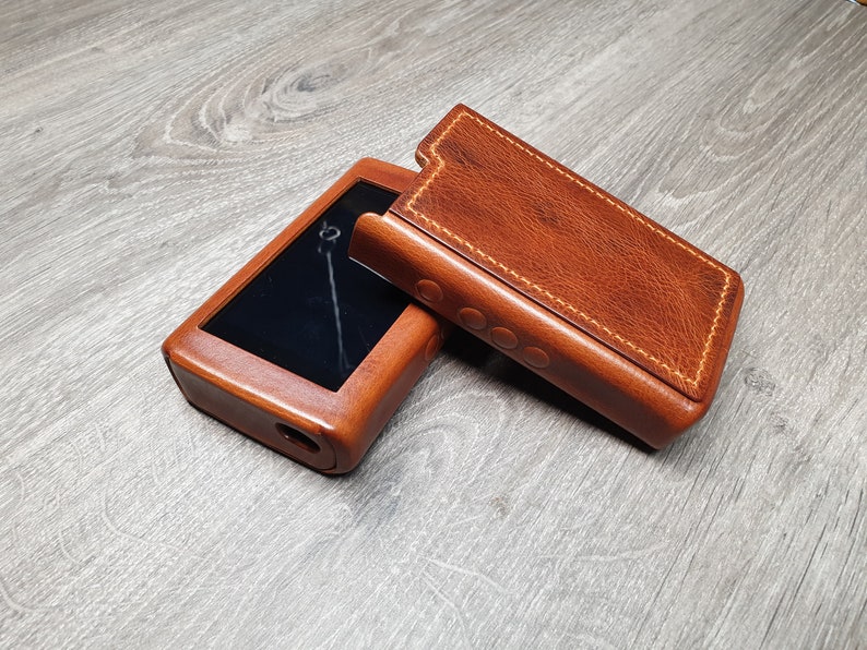 Puede incluir: Una funda y tapa de cuero marr&oacute;n para un reproductor de m&uacute;sica port&aacute;til. La funda est&aacute; abierta, revelando la pantalla del reproductor. El cuero tiene un tono rico y c&aacute;lido, con costuras visibles. El reproductor tiene una pantalla negra y un puerto circular.