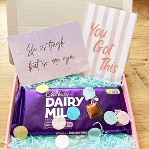 May include: A gift box containing a Cadbury Dairy Milk chocolate bar, two cards with motivational messages, and confetti. The cards say "Life is tough but so are you" and "You Got This". The box is pink and filled with blue paper.