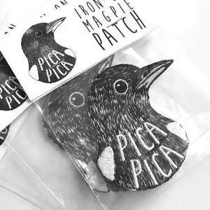 Può includere: Due toppe termoadesive in bianco e nero di gazze con il testo "Pica Pica" su ciascuna. Le toppe sono su uno sfondo bianco.