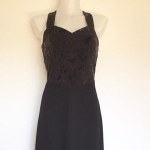 May include: A black formal gown with a halter neckline. The bodice features a textured, dark floral pattern, transitioning to a smooth, floor-length skirt. The dress is displayed on a mannequin.