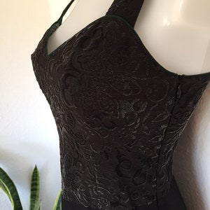 May include: Black evening dress with a textured bodice featuring an intricate floral pattern. The dress has a halter neckline and a flowing skirt. A zipper runs along the side. The dress is displayed on a mannequin.