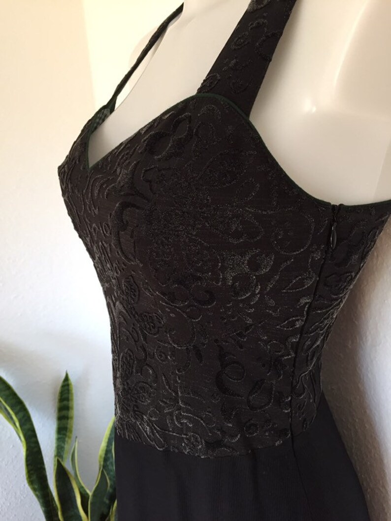 May include: Black evening dress with a textured bodice featuring an intricate floral pattern. The dress has a halter neckline and a flowing skirt. A zipper runs along the side. The dress is displayed on a mannequin.