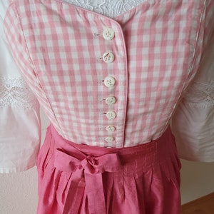 May include: A pink and white gingham bodice with white buttons and a pink skirt with a bow. The bodice has a white lace trim.