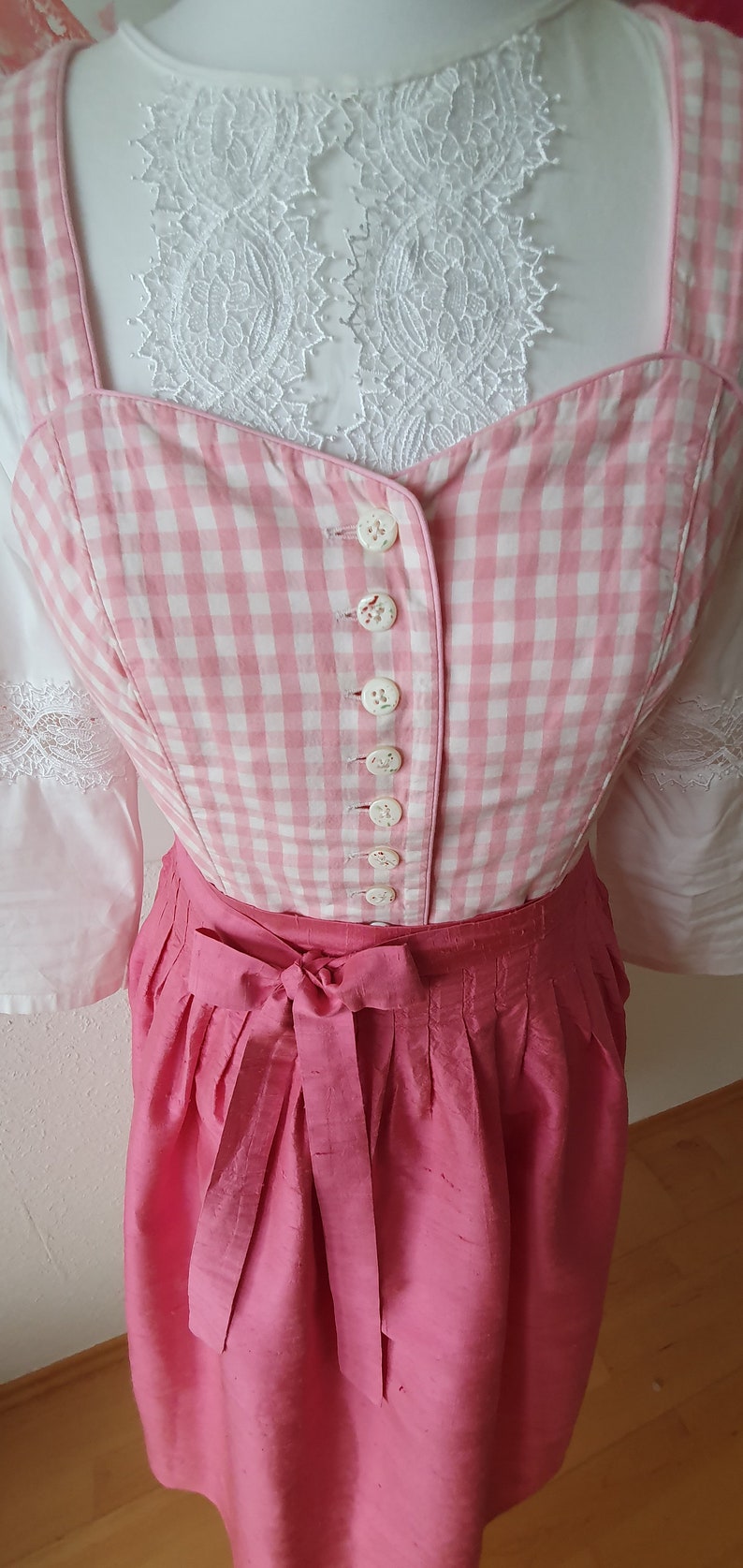 May include: A pink and white gingham bodice with white buttons and a pink skirt with a bow. The bodice has a white lace trim.
