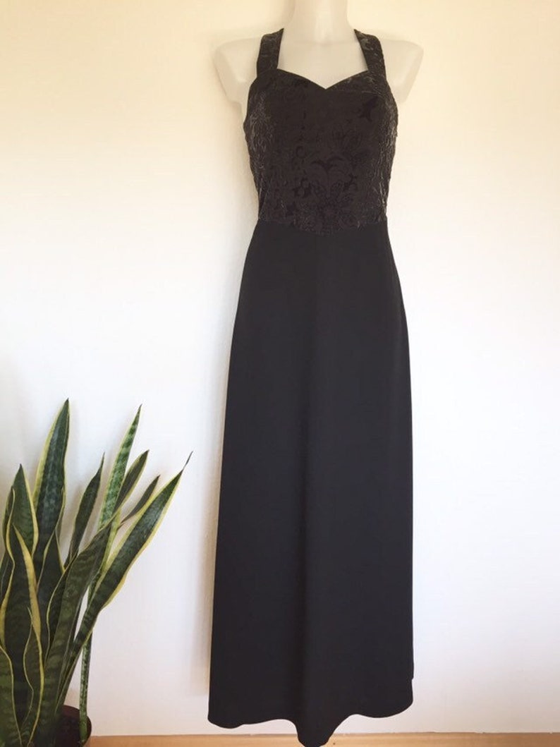 May include: A black formal gown with a halter neckline. The bodice features a textured, dark floral pattern, transitioning to a smooth, floor-length skirt. The dress is displayed on a mannequin.