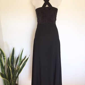 May include: A black evening gown with a halter neckline and a floor-length skirt. The bodice features a dark, textured pattern, and the dress is displayed on a mannequin against a white wall. A potted plant is visible in the corner.
