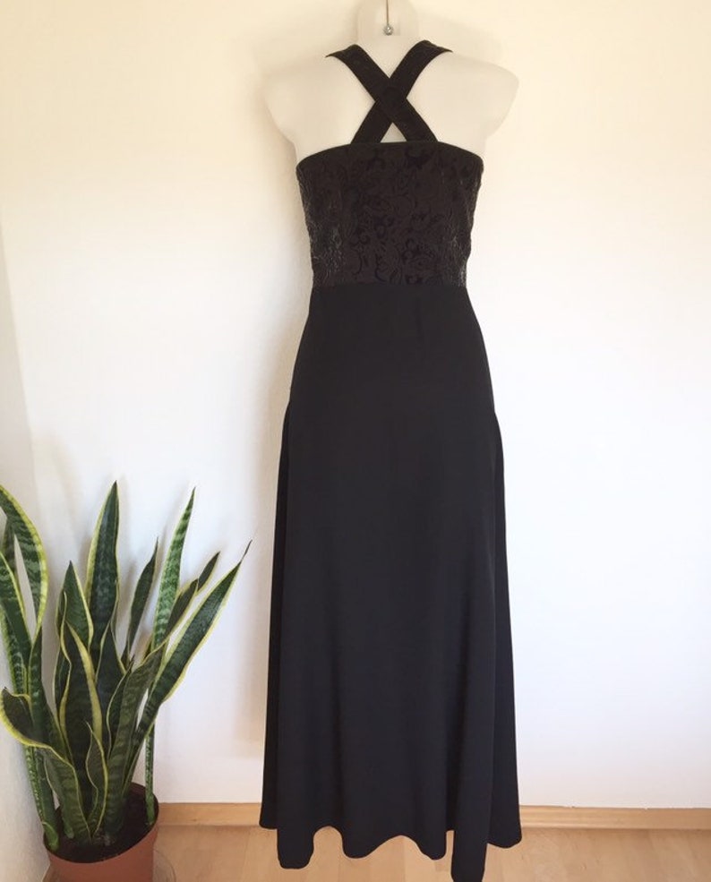 May include: A black evening gown with a halter neckline and a floor-length skirt. The bodice features a dark, textured pattern, and the dress is displayed on a mannequin against a white wall. A potted plant is visible in the corner.