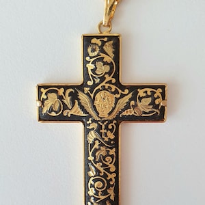 Könnte beinhalten: Ein goldfarbener Kreuzanhänger mit aufwendigen schwarzen und goldenen floralen und figürlichen Designs. Das Kreuz hängt an einer goldenen Kette. Der Anhänger zeigt ein detailliertes Gesicht mit Flügeln und verzierten Mustern.