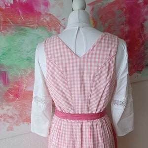 May include: Pink and white gingham dirndl dress with a pink ribbon sash. The dress has a v-neck and is sleeveless. The dress is worn on a white mannequin.