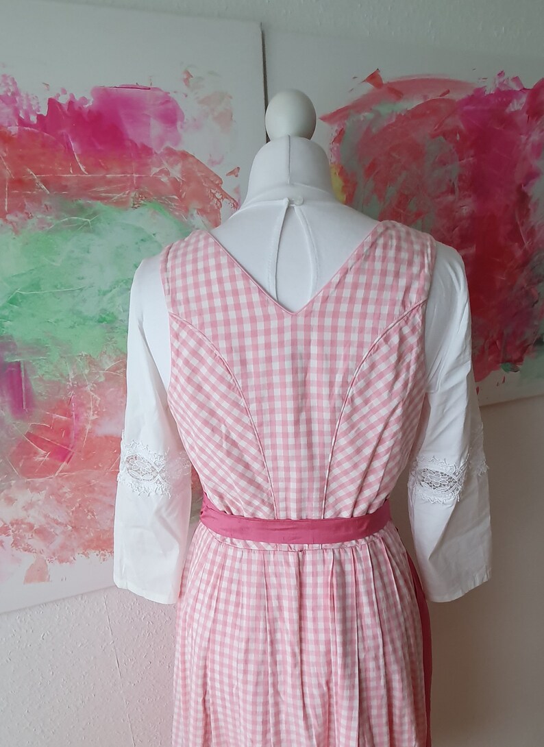 May include: Pink and white gingham dirndl dress with a pink ribbon sash. The dress has a v-neck and is sleeveless. The dress is worn on a white mannequin.