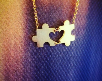 May include: A gold-colored necklace featuring two puzzle piece pendants connected by a heart-shaped cutout. The pendants are linked by a delicate chain, creating a symbol of unity and connection. The necklace is displayed against a textured background.