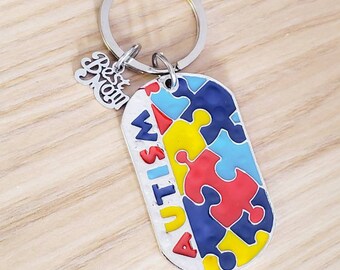 May include: A silver keychain with a rectangular pendant featuring a colorful puzzle design. The word "AUTISM" is vertically displayed. A small charm reads "Best Mom". The keychain is on a light wood surface.