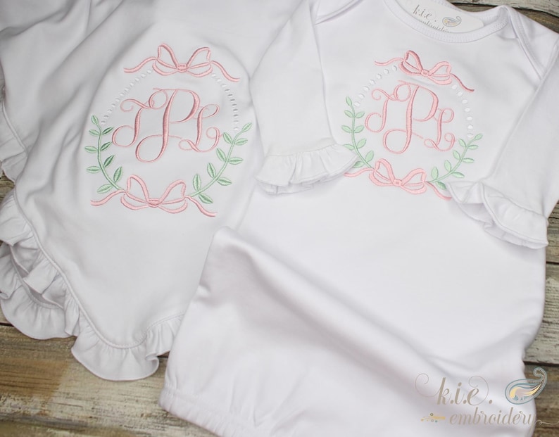 Op de afbeelding: Witte babyjurk en bijpassend dekentje met geborduurd monogram in roze en groen in een kransontwerp.