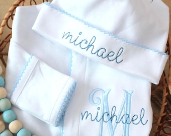 May include: White baby blanket, hat, and burp cloth set with light blue embroidered details. The name "michael" is embroidered on the hat and blanket. The blanket also features a large embroidered "M". The set is displayed in a woven basket.