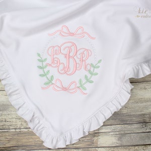 Op de afbeelding: Wit babydekentje met een gerimpelde rand en een geborduurd monogram in het midden. Het monogram is omringd door een krans van groene bladeren en roze strikken.